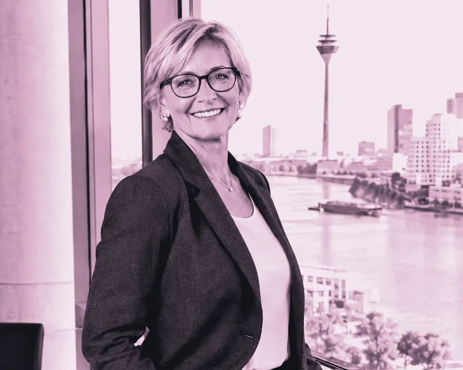 Frau mit kurzen blonden Haaren und Brille im anthrazitfarbenen Hosenanzug lächelt am Fenster eines Besprechungsraums mit Blick auf den Rhein